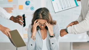 A business woman looking stressed, people surround her with papers and graphs.