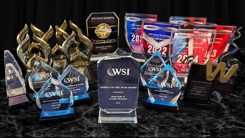 Many different award trophies staged on a dark marble counter in front of a dark curtain.